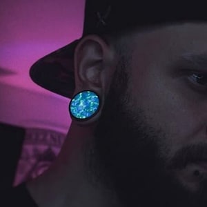 May include: A man with a beard and a black baseball cap is wearing a blue and green ear gauge. He has a nose ring with three silver rings.