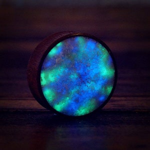 May include: A round wooden ear gauge with a blue and green glow-in-the-dark resin inlay. The resin has a speckled pattern.