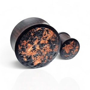 May include: Two dark brown wooden ear plugs with a textured black and copper-colored design on the front. The plugs are cylindrical with a flared edge. The background is white.