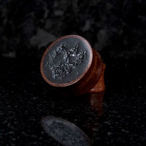 May include: A pair of wooden ear gauges with a black resin inlay featuring a unique design. The gauges are a dark brown color and have a smooth, polished finish.