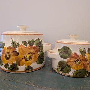May include: Two off-white ceramic lidded pots with side handles, featuring hand-painted yellow and orange flowers with green leaves. The pots have a smooth, glossy finish and orange-brown trim along the rims and lids, showcasing a floral design.