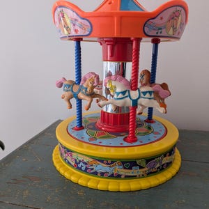 May include: A colorful plastic carousel toy featuring a red roof with blue accents, a central mirrored pillar, and four small horses with pink manes. The base is yellow with a decorative band showing illustrations of horses, musical notes, and hearts.