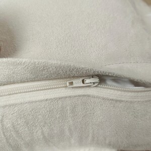 May include: Close-up of a cream-colored, plush fabric with a white zipper. The zipper is partially open, revealing the interior of the fabric. The fabric has a soft, textured appearance.