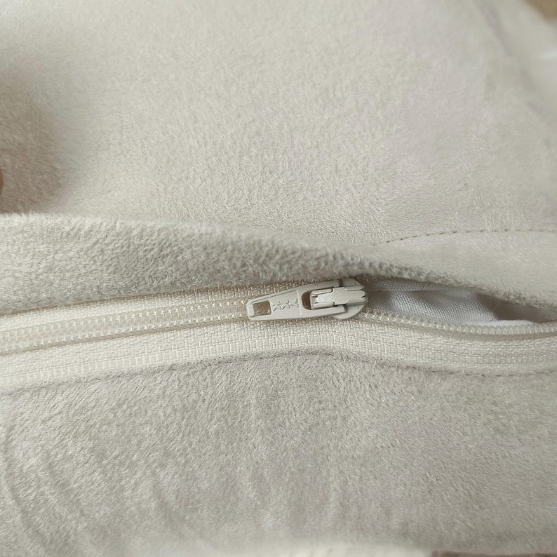 May include: Close-up of a cream-colored, plush fabric with a white zipper. The zipper is partially open, revealing the interior of the fabric. The fabric has a soft, textured appearance.