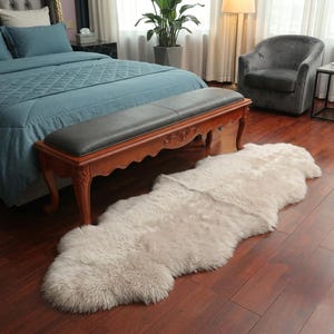 May include: A light beige sheepskin rug on a dark wood floor. The rug is irregularly shaped and has a soft, fluffy texture. A wooden bench with a dark gray cushion is behind the rug. The room has a bed with blue bedding and a gray armchair.