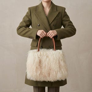 Pode incluir: Um blazer de lã verde-oliva com abotoamento duplo, lapelas e botões escuros, combinado com uma bolsa retangular de pelo sintético longo e felpudo na cor creme, com duas alças superiores arredondadas em couro marrom.