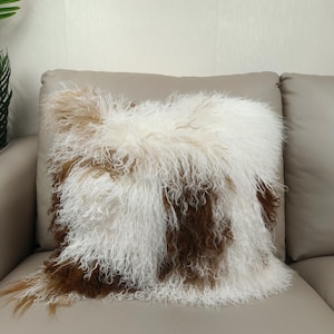 May include: A square decorative pillow with a shaggy, textured cover. The pillow features a mix of white, brown, and tan fur-like material, creating a soft and inviting appearance. The pillow is placed on a neutral-toned sofa.