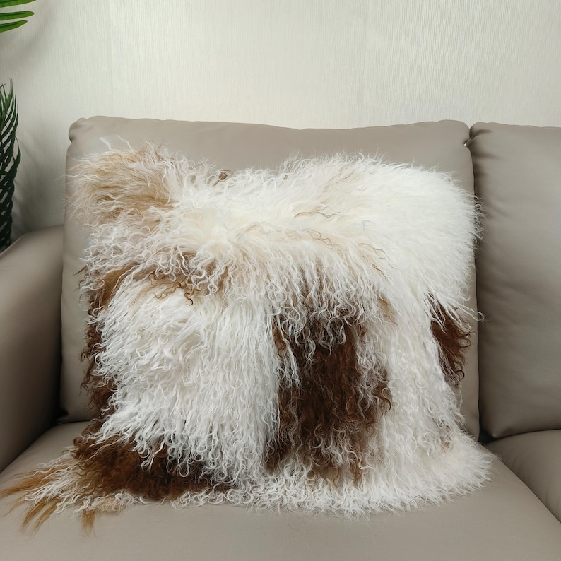 May include: A square decorative pillow with a shaggy, textured cover. The pillow features a mix of white, brown, and tan fur-like material, creating a soft and inviting appearance. The pillow is placed on a neutral-toned sofa.