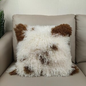 May include: Square decorative pillow with a shaggy, curly texture. The pillow features a white base with brown patches, resembling a sheep or dog. The pillow is placed on a beige sofa.