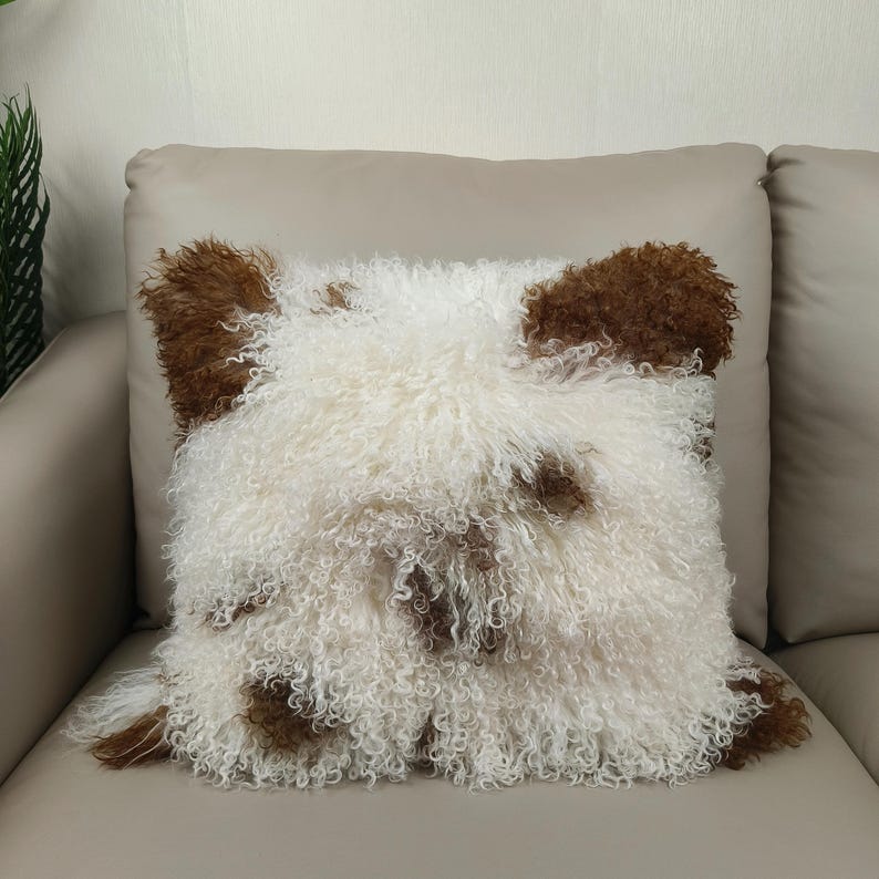 May include: Square decorative pillow with a shaggy, curly texture. The pillow features a white base with brown patches, resembling a sheep or dog. The pillow is placed on a beige sofa.