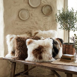 May include: A collection of square, shaggy throw pillows in shades of brown, white, and black, arranged on a rustic wooden bench. The pillows have a textured, fur-like appearance. Decorative plates hang on the textured wall in the background.