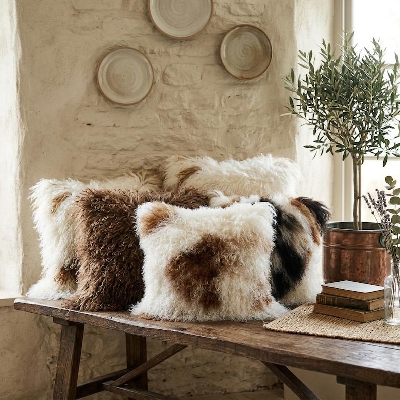 May include: A collection of square, shaggy throw pillows in shades of brown, white, and black, arranged on a rustic wooden bench. The pillows have a textured, fur-like appearance. Decorative plates hang on the textured wall in the background.