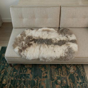 May include: A plush, irregular-shaped sheepskin rug featuring a natural color palette of cream, beige, and soft brown tones. The thick, long-pile wool has a variegated, mottled appearance with darker patches distributed across the textured surface.