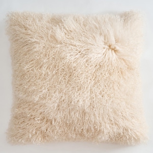 May include: A square throw pillow covered in long, curly, cream-colored fur. The texture is dense and shaggy, featuring a distinct swirl pattern near the upper right corner where the fibers converge. The pillow has a soft, voluminous appearance.