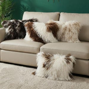 Puede incluir: Una colección de cojines decorativos con superficies peludas y texturizadas. Los cojines presentan una mezcla de colores, incluyendo marrón oscuro, marrón claro y blanco. Están colocados sobre un sofá beige y una alfombra de color claro.