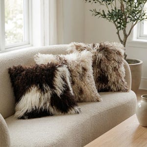 May include: Three square throw cushions with long, shaggy, curly fur textures. The leftmost cushion features a high-contrast pattern of dark brown and cream white. The middle and right cushions display variegated shades of tan, brown, and cream.
