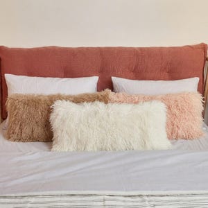 May include: A rectangular, tufted, terracotta-colored headboard sits behind two white rectangular pillows. In front are three shaggy, faux-fur rectangular pillows in shades of light brown, cream, and soft pink, arranged horizontally on a bed.