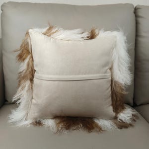 May include: A beige decorative pillow with a shaggy, fur-like trim in shades of brown and white. The pillow has a soft, suede-like texture and is placed on a neutral-toned sofa. The pillow's design adds a touch of texture and warmth.