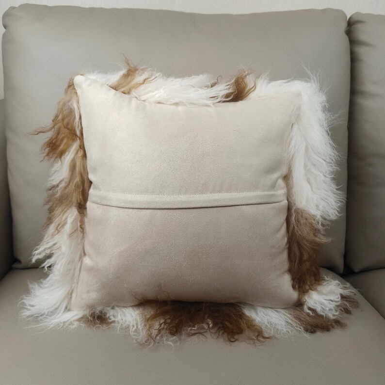 May include: A beige decorative pillow with a shaggy, fur-like trim in shades of brown and white. The pillow has a soft, suede-like texture and is placed on a neutral-toned sofa. The pillow's design adds a touch of texture and warmth.