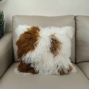 May include: A square decorative pillow with a shaggy, textured surface. The pillow features a pattern of white and brown fur, creating a unique and cozy aesthetic. The pillow is placed on a neutral-toned sofa.