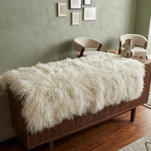 May include: A cream-coloured, shaggy sheepskin rug draped over a dark wood cabinet. The rug has long, curly fibres. The cabinet has a mid-century modern design with four doors and angled legs. Two beige armchairs and wall art are in the background.