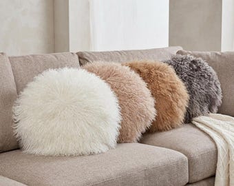 May include: A set of four round throw pillows with long, shaggy faux fur texture. The pillows are arranged in a row, featuring colors of cream, light beige, medium tan, and dark charcoal gray, creating a soft and layered appearance.