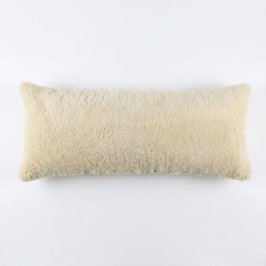 Puede incluir: Un cojín rectangular con una textura gruesa de imitación de piel de oveja en color crema. El cojín tiene un aspecto suave y afelpado con una superficie de pelo denso y rizado que cubre toda la parte frontal de la forma rectangular.