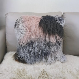 May include: A square decorative pillow with a shaggy, textured surface. The pillow features a color block design with sections of pink, gray, and black. The pillow is placed on a white surface.