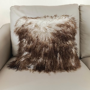 May include: A decorative square pillow with a shaggy, textured surface. The pillow's design features a combination of brown and white fur, creating a unique and cozy aesthetic. The pillow is placed on a light-colored sofa.