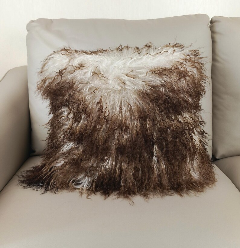 May include: A decorative square pillow with a shaggy, textured surface. The pillow's design features a combination of brown and white fur, creating a unique and cozy aesthetic. The pillow is placed on a light-colored sofa.