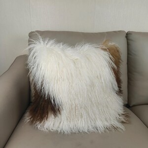 May include: A square decorative pillow with a shaggy, textured surface. The pillow features a mix of white, brown, and tan fur, creating a layered effect. The pillow is placed on a neutral-toned sofa.