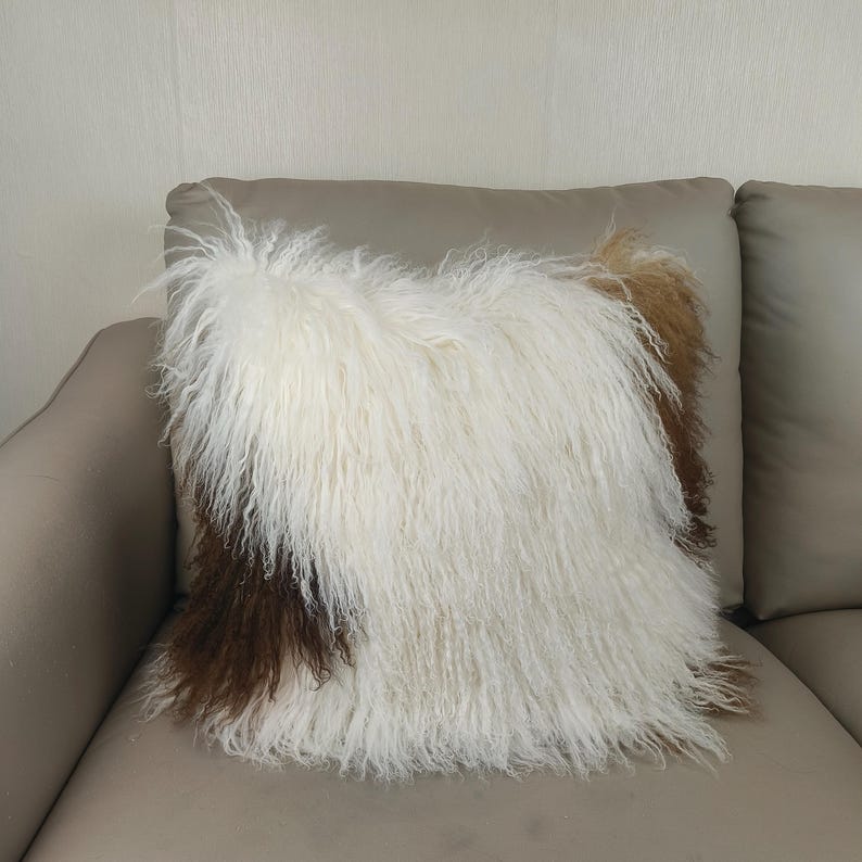 May include: A square decorative pillow with a shaggy, textured surface. The pillow features a mix of white, brown, and tan fur, creating a layered effect. The pillow is placed on a neutral-toned sofa.