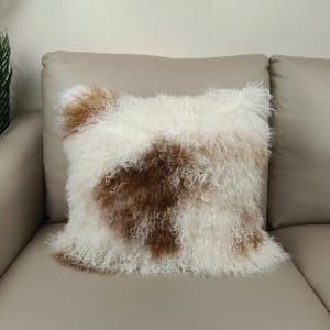 May include: Square decorative pillow with a shaggy, textured surface. The pillow features a pattern of white, brown, and tan fur. The pillow is placed on a beige sofa, creating a cozy and inviting aesthetic.