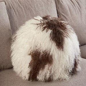 Könnte beinhalten: Ein rundes, flauschiges Kissen mit weißem und braunem Fell. Das Kissen liegt auf einem hellbraunen Sofa.