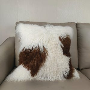 May include: Square decorative pillow with a shaggy texture. The pillow features a pattern of white and brown fur, creating a cozy and stylish accent for home decor. The pillow is placed on a neutral-toned sofa.