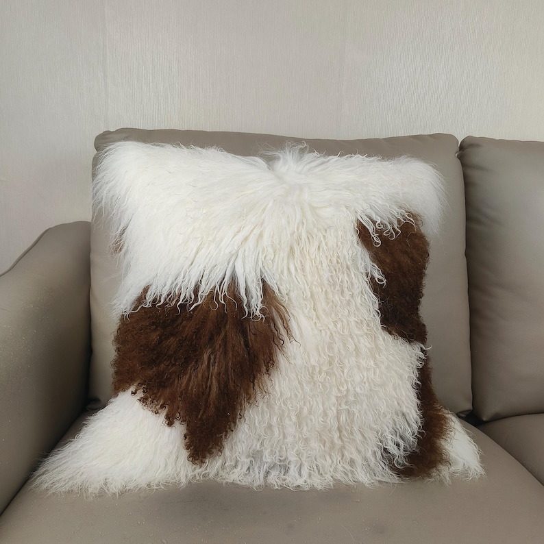 May include: Square decorative pillow with a shaggy texture. The pillow features a pattern of white and brown fur, creating a cozy and stylish accent for home decor. The pillow is placed on a neutral-toned sofa.
