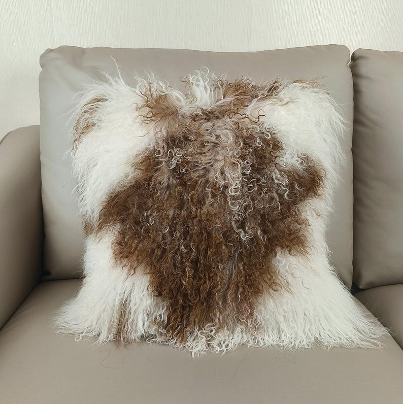 May include: Square decorative pillow with a brown and white fur design. The pillow has a soft, textured appearance, with the brown fur forming a central pattern against a white background. The pillow is placed on a neutral-toned sofa.