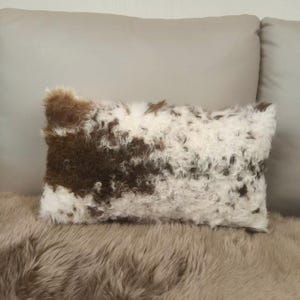 Puede incluir: Un cojín decorativo rectangular con una superficie peluda y texturizada. El cojín presenta un patrón de piel marrón y blanca, creando una estética acogedora y atractiva. El cojín está sobre una superficie peluda.