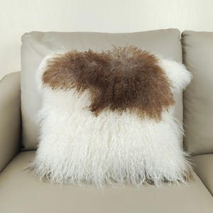 May include: A square decorative pillow with a white and brown fur design. The pillow has a fluffy, textured appearance, with a patch of brown fur in the center. The pillow is placed on a light-colored sofa.