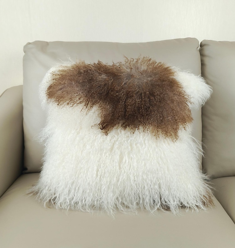 May include: A square decorative pillow with a white and brown fur design. The pillow has a fluffy, textured appearance, with a patch of brown fur in the center. The pillow is placed on a light-colored sofa.