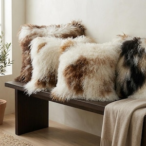 May include: Four decorative throw pillows with shaggy, textured covers in shades of brown, white, and black rest on a dark wooden bench. The pillows have a fluffy, fur-like appearance, adding a cozy and inviting touch to the space.