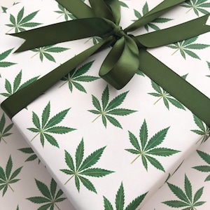 Puede incluir: Papel de regalo blanco con un patrón repetido de hojas verdes de marihuana. Una cinta de satén verde está atada en un lazo en la parte superior del papel de regalo.