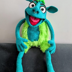 May include: A handmade teal and green puppet with large eyes, a red tongue, and a nose ring. The puppet has a furry green vest and shorts, with long teal arms and legs. It is sitting on a dark surface.