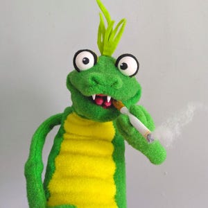 May include: A plush green alligator puppet with a yellow belly and a tuft of green hair. The alligator is smoking a cigarette, with smoke rising from it. The puppet has large, googly eyes and a toothy grin.
