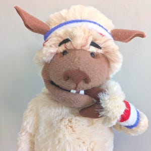 Peut inclure: Une peluche mouton avec un corps crème, des oreilles marron et un visage marron. Le mouton porte un bandeau et un bracelet bleu, blanc et rouge. Le mouton sourit et montre deux dents.