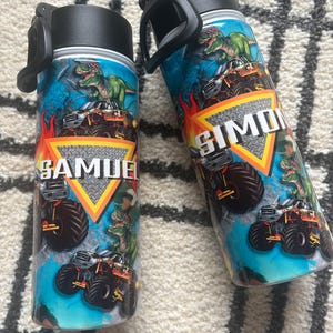 May include: Two personalized water bottles with black lids and handles. The bottles feature a design with a green dinosaur, a monster truck, and the names "SAMUEL" and "SIMON" in a bold font against a blue and orange background.