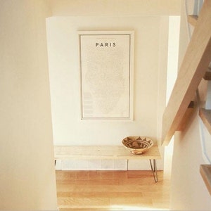 May include: A white wooden bench with black hairpin legs sits in a hallway with a framed map of Paris on the wall above it. A woven basket sits on the bench.
