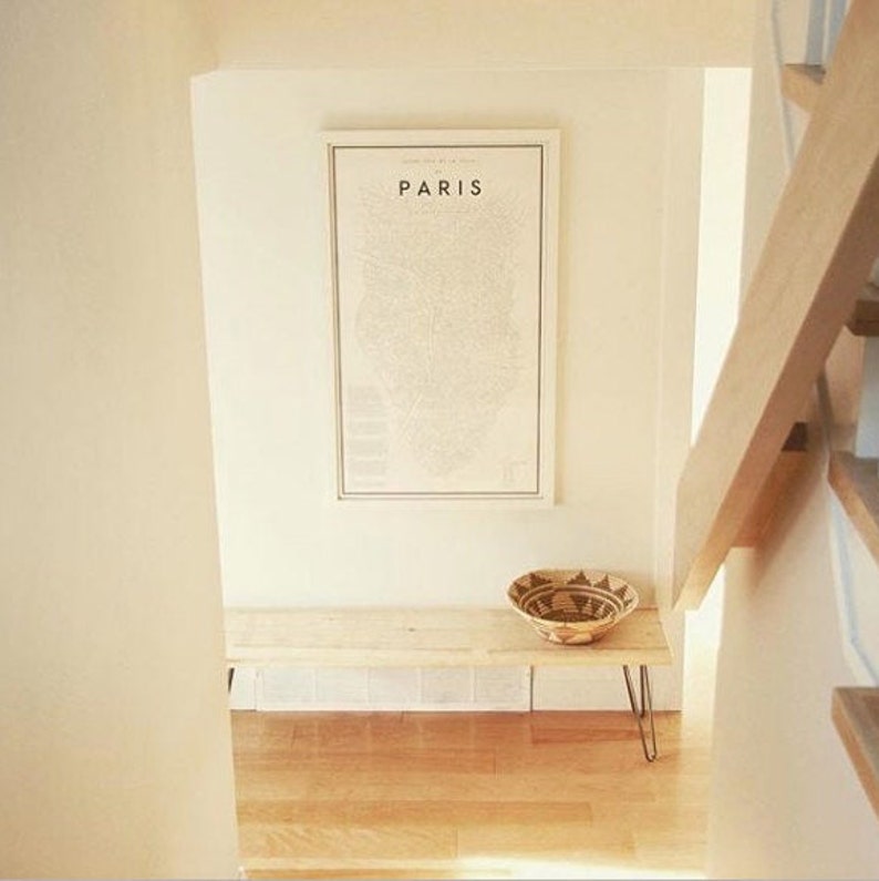 May include: A white wooden bench with black hairpin legs sits in a hallway with a framed map of Paris on the wall above it. A woven basket sits on the bench.