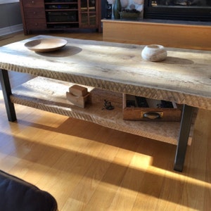 Pu&ograve; includere: Un tavolino da caff&egrave; in legno rustico con un piano in legno vivo e un ripiano inferiore. Il tavolo ha gambe in metallo nero e presenta una ciotola di legno, una piccola scatola di legno e un oggetto decorativo sullo scaffale.