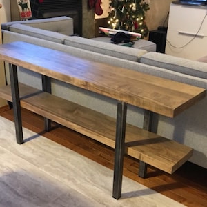 Pu&ograve; includere: Un tavolo console lungo in legno con due ripiani e gambe in metallo nero. Il tavolo &egrave; realizzato in legno chiaro e ha una finitura naturale. Il tavolo &egrave; in un soggiorno, davanti a un divano.